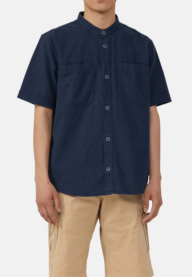 Marineblauw overhemd met korte mouwen en staande kraag, voor sluiting met knopen, twee borstzakken en een zachte katoentextuur. Gecombineerd met beige cargoshorts.