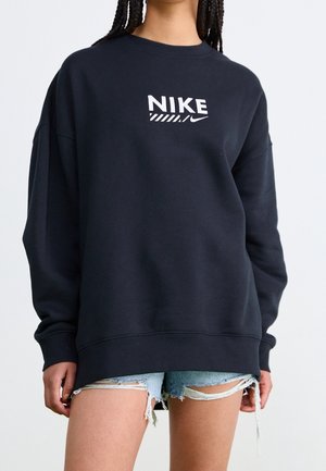 Osoba ubrana w czarną bluzę Nike z białym logo i jasno-niebieskie podarte szorty dżinsowe, stojąca na jednolitym tle.