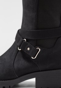 Bottine en suède noir avec talon épais, sangle décorative et détail de boucle en métal argenté sur le côté.