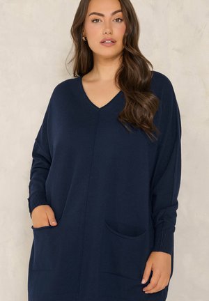 Vrouw met lang bruin haar die een marineblauwe V-hals trui-jurk met zakken draagt, staande voor een licht beige achtergrond.