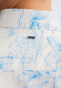 Witte shirt met blauwe bloemenprint, met een button-down kraag en een klein zilveren logo-label bij de kraag aan de achterkant.
