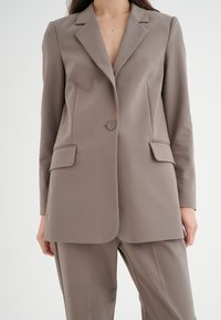 Blazer marrone chiaro in un tessuto liscio, con chiusura a singolo bottone, due tasche frontali e taglio su misura.