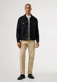 Tamno traper jakna s prednjim džepovima preko prugaste košulje, uparena s kaki cargo hlačama i crnim cipelama. Ima klasičan kroj i dizajn.