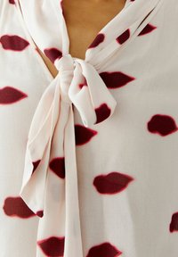 Leichtes, seidiges Blusenshirt mit Schluppe und einem Muster aus roten Lippenformen auf hellem Hintergrund. Der Stoff hat eine glatte Textur.