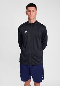 Zwarte sportjack met een ritssluiting aan de voorkant, staande kraag en wit logo. Gecombineerd met marineblauwe shorts, gemaakt van lichtgewicht stof.