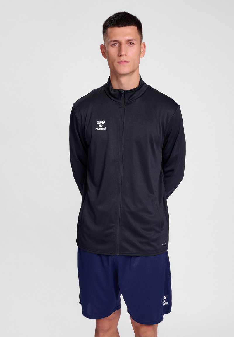 Zwarte sportjack met een ritssluiting aan de voorkant, staande kraag en wit logo. Gecombineerd met marineblauwe shorts, gemaakt van lichtgewicht stof.