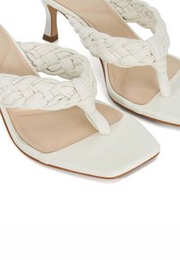 White heeled sandals feature braided straps, square toes, and a smooth beige insole. The heel has a contrasting brown base.