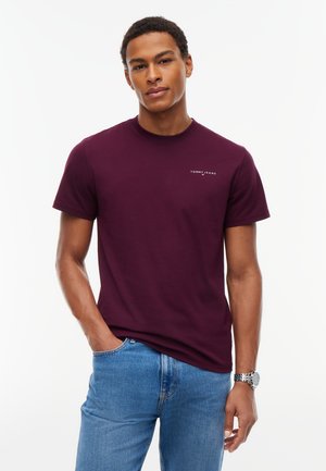 Giovane uomo con capelli corti e ricci che indossa una t-shirt Tommy Jeans color bordeaux e jeans blu, in piedi con una mano in tasca su sfondo bianco.