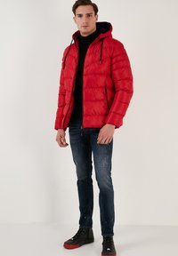 Rød pufferjakke med hætte, lynlåslukning foran og to sidelommer. Bæres over en sort sweater, parret med mørke jeans og sorte sneakers.