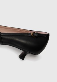 Scarpa nera in pelle con tacco kitten e punta rotonda, caratterizzata da un piccolo dettaglio con fibbia dorata sul lato e una superficie liscia.