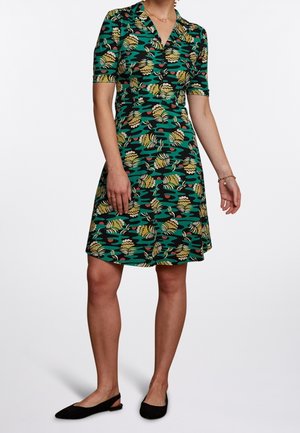 Woman wearing a green knee-length dress with short sleeves and a yellow and black floral pattern, paired with black flat shoes.