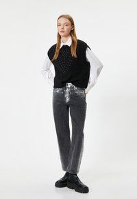 Schwarze gestrickte ärmellose Weste mit strukturiertem Muster und silbernen Verzierungen, kombiniert mit dunkelgrauen Jeans mit silbernen Akzenten und schwarzen Plateaustiefeln.