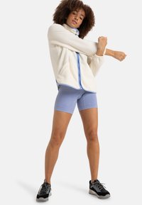 Giacca di pile crema con cerniera e dettagli blu, abbinata a pantaloni corti leggeri a coste blu chiaro. Scarpe da ginnastica nere e grigie completano l'outfit.
