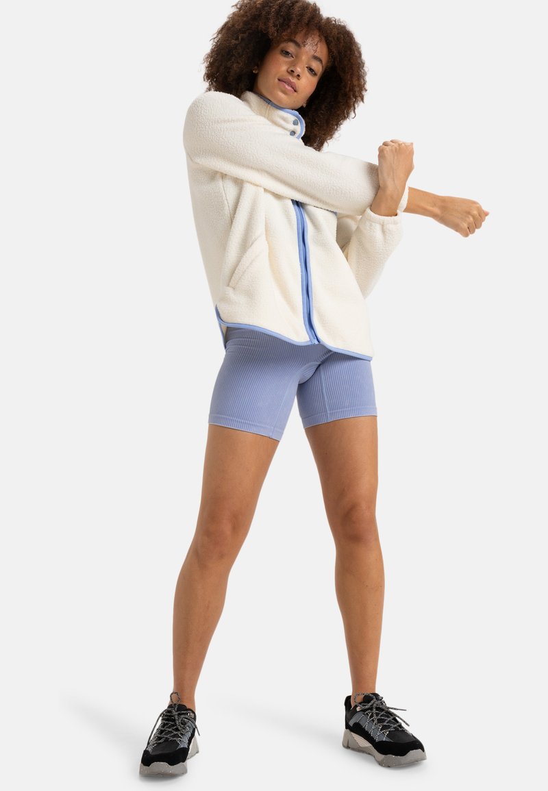 Kermansin fleece takki sinisellä vetoketjulla ja yksityiskohdilla, yhdistettynä ribattyihin vaaleansinisiin shortseihin. Mustat ja harmaat urheilukengät viimeistelevät asun.