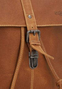 Cinghia di zaino in pelle marrone con fibbia metallica e dettagli cuciti. La vista ravvicinata mostra la texture e i dettagli di chiusura sulla pelle.