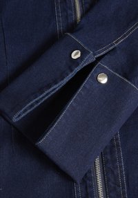 Jeansjacka i mörkblå färg, med silverknappar, kontrasterande sömmar och en synlig dragkedja framtill.