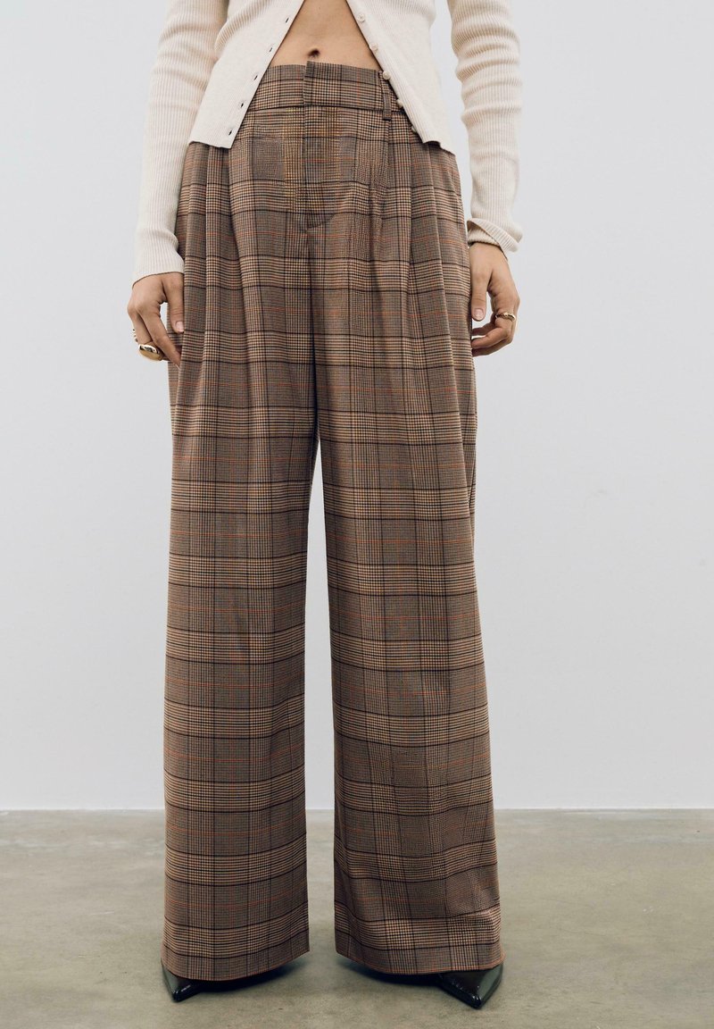Pantalons larges à carreaux marron avec une taille haute, présentant des plis et un tissu texturé. Associés à un cardigan de couleur claire et des talons.