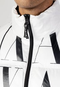 Giacca imbottita bianca con colletto alto, zip nera e un prominente motivo geometrico nero. Il materiale sembra lucido e strutturato.