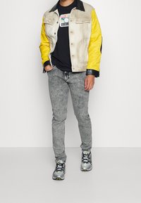 Chaqueta con bloques de color con mangas amarillas, cuerpo beige y cuello negro; camiseta negra; jeans grises con patrón; zapatillas grises con detalles.