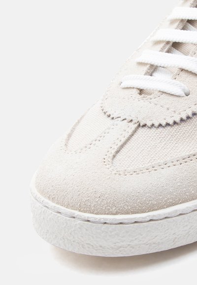 Κοντινό πλάνο στη μύτη ενός μπεζ αθλητικού παπουτσιού από σουέτ και δίχτυ, με λευκή υφή στη σόλα και λευκά κορδόνια, σε απλό λευκό φόντο.