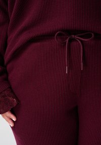 Close-up van een persoon die bordeauxrode geribbelde gebreide broek en bijpassend top draagt, met een gestrikt koord in de taille en kantafwerking op de mouwen.