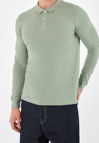 Langsleeve poloshirt in gedempt groen katoen, voorzien van een drieknopenlijst, geribde manchetten en een zachte, gladde textuur. Gepaard met donkere jeans.