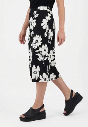 Schwarzer Midirock mit großem weißem Blumenmuster, getragen mit schwarzen offenen Keilsandalen, zeigt seitlich und teilweise den unteren Körper.