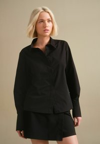 Zwarte button-up shirt met lange mouwen, puntige kraag en een gestructureerde pasvorm, gecombineerd met een bijpassende zwarte rok. Gladde textuur van de stof.