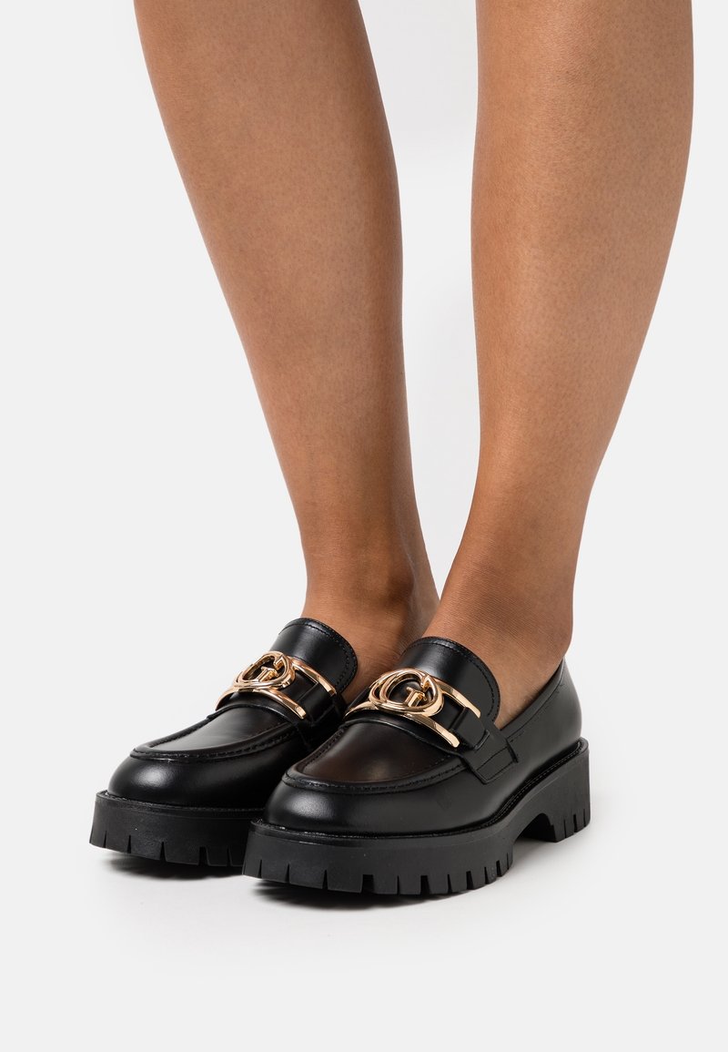 Mocassins épais en cuir noir avec semelles épaisses et boucles décoratives dorées portés pieds nus sur fond blanc.