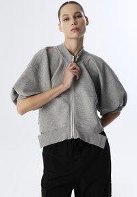 Grijze zip-up sweatshirt met brede, korte mouwen en ribbelrand. Gladde textuur met minimale naden, gedragen over losse zwarte broek.