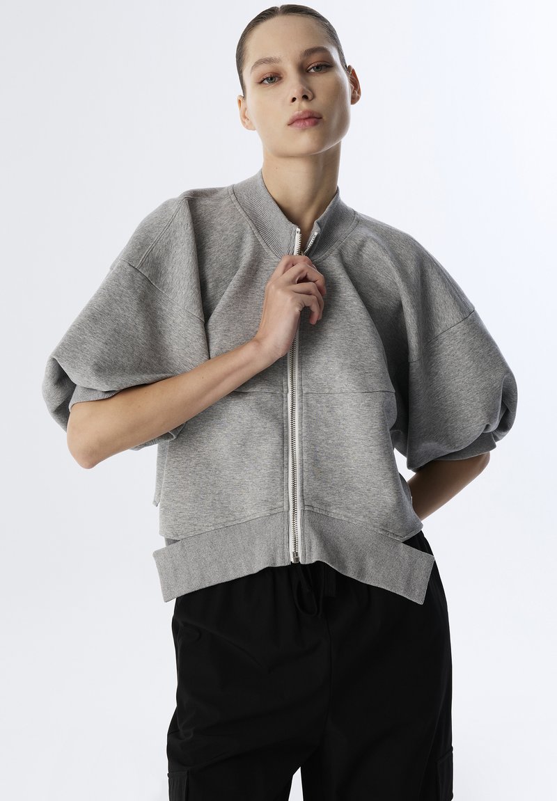 Grijze zip-up sweatshirt met brede, korte mouwen en ribbelrand. Gladde textuur met minimale naden, gedragen over losse zwarte broek.
