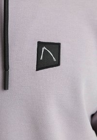 Light purple hoodie featuring a black rectangular patch with white geometric stitching. Drawstrings are black with a plastic tip.