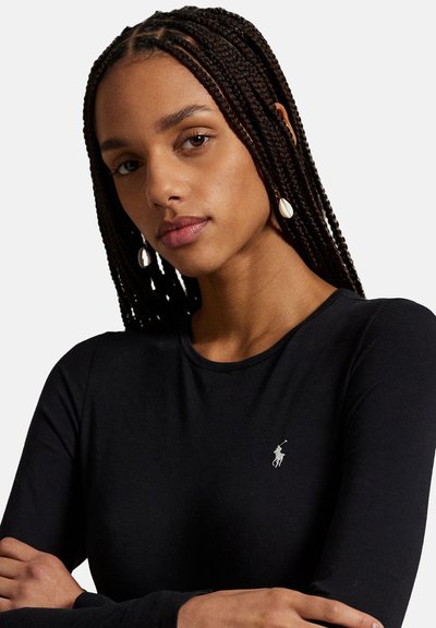 Femme aux longs cheveux tressés portant des boucles d'oreilles en coquillage et une chemise à manches longues noire avec un petit logo Polo blanc sur la poitrine.