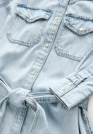 Camicia di denim azzurro chiaro con due tasche sul petto, bottoni a pressione, maniche arrotolate e una cintura in tessuto abbinata legata in vita.