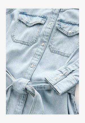 Camisa de denim azul claro con dos bolsillos en el pecho, botones a presión, mangas enrolladas y un cinturón de tela a juego atado en la cintura.