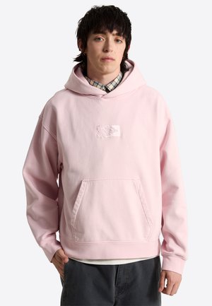 Joven con cabello oscuro que lleva una sudadera con capucha rosa claro sobre una camisa de cuadros abotonada y pantalones oscuros, de pie con una mano en el bolsillo.