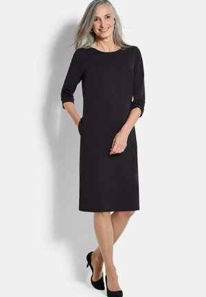 Lächelnde reife Frau mit grauen Haaren in einem schwarzen knielangen Kleid und schwarzen Schuhen mit Absatz, die mit einer Hand in der Tasche vor weißem Hintergrund steht.