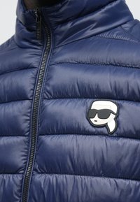 Nærbilde av en marineblå quiltet jakke med glidelås foran og en liten lapp med en person som har på seg solbriller på brystet.