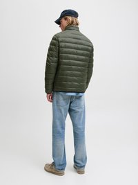 Jachetă umplută de culoare verde olivescu, cu guler înalt, asortată cu blugi ușor albaștri. Persoana poartă o șapcă bleumarin și pantofi bej.