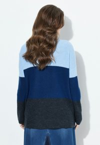 Eine Frau mit langen, gewellten braunen Haaren trägt einen Farbblock-Pullover in Hellblau, Marineblau und Dunkelgrau, kombiniert mit einem blauen Denim-Rock, während sie vor einem weißen Hintergrund steht.