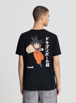 Młody mężczyzna w czarnej koszulce z postacią Goku z Dragon Ball w pomarańczowym stroju, wyprowadzający cios w przód, z japońskim napisem na plecach.