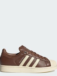 Καφέ δερμάτινο sneaker Adidas Superstar με κρεμ ρίγες, κορδόνια, λαστιχένια μύτη και σόλα, ορατό από δεξιά πλευρά.