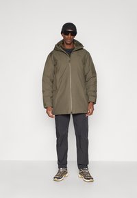 Giacca impermeabile verde oliva con chiusura a zip, cappuccio e tasche. Abbinata a pantaloni scuri e scarpe da trekking marroni. Berretto nero e occhiali da sole.