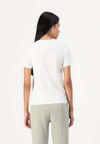 T-shirt bianco in cotone con maniche corte e scollo rotondo, visto da dietro. Abbinato a pantaloni verde chiaro. Tessuto liscio.