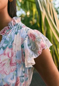 Gėlių motyvu palaidinė su švelniomis pastelinėmis spalvomis, rožinėmis ir žaliomis gėlėmis ant šviesiai mėlynos medžiagos. Raukiniai ant rankovių ir sugrupuota iškirptės detalė.