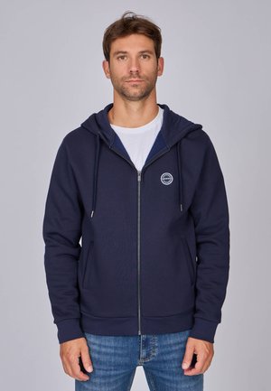 Sweat-shirt zippé bleu marine avec capuche à cordon, poches avant et patch logo. Fabriqué en tissu doux avec des poignets et un ourlet côtelés. Porté sur une chemise blanche.