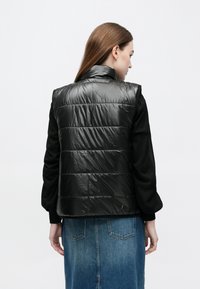 Γυναίκα με μακριά καστανά μαλλιά που φοράει μια γυαλιστερή μαύρη γιλέκο με ραφή πάνω από ένα μαύρο πουλόβερ και μια μπλε τζιν φούστα, με την πλάτη στραμμένη σε λευκό φόντο.