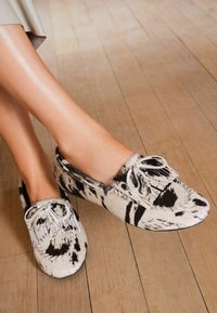 Lábak fekete-fehér tehénbőr mokaszín lófaszban, fráncsos és masnis díszítéssel, egy fa padlón.