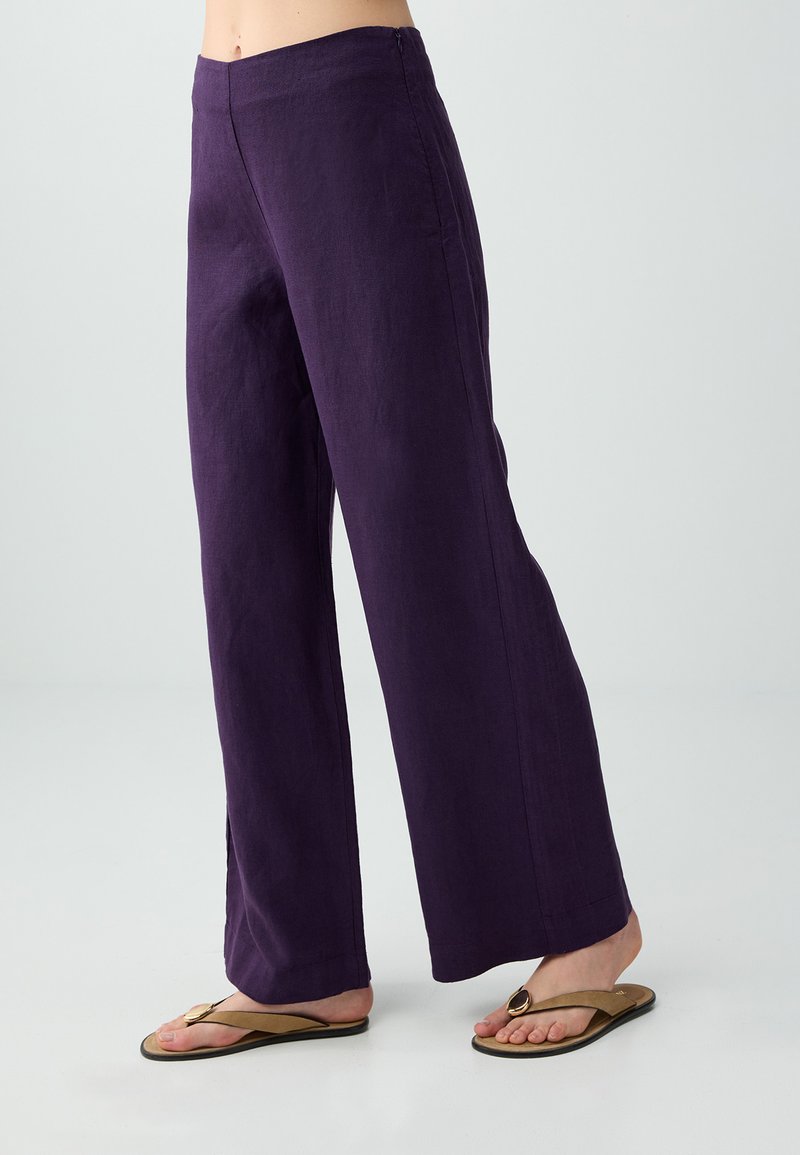 Partie inférieure d'une personne portant un pantalon ample violet foncé et des tongs beige avec un détail d'anneau métallique, debout sur un sol blanc.