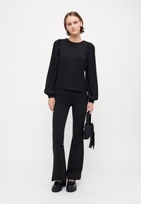 Black ribbed sweater with puffed long sleeves, paired with black flared pants and glossy black shoes. Holds a small black handbag.